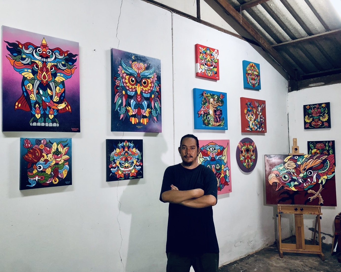 ชายเสื้อดำยืนกอดอกท่ามกลางภาพสัตว์สีสดบนผนังสีขาว ด้านขวามีขาตั้งไม้รองรับภาพสัตว์ตาโต