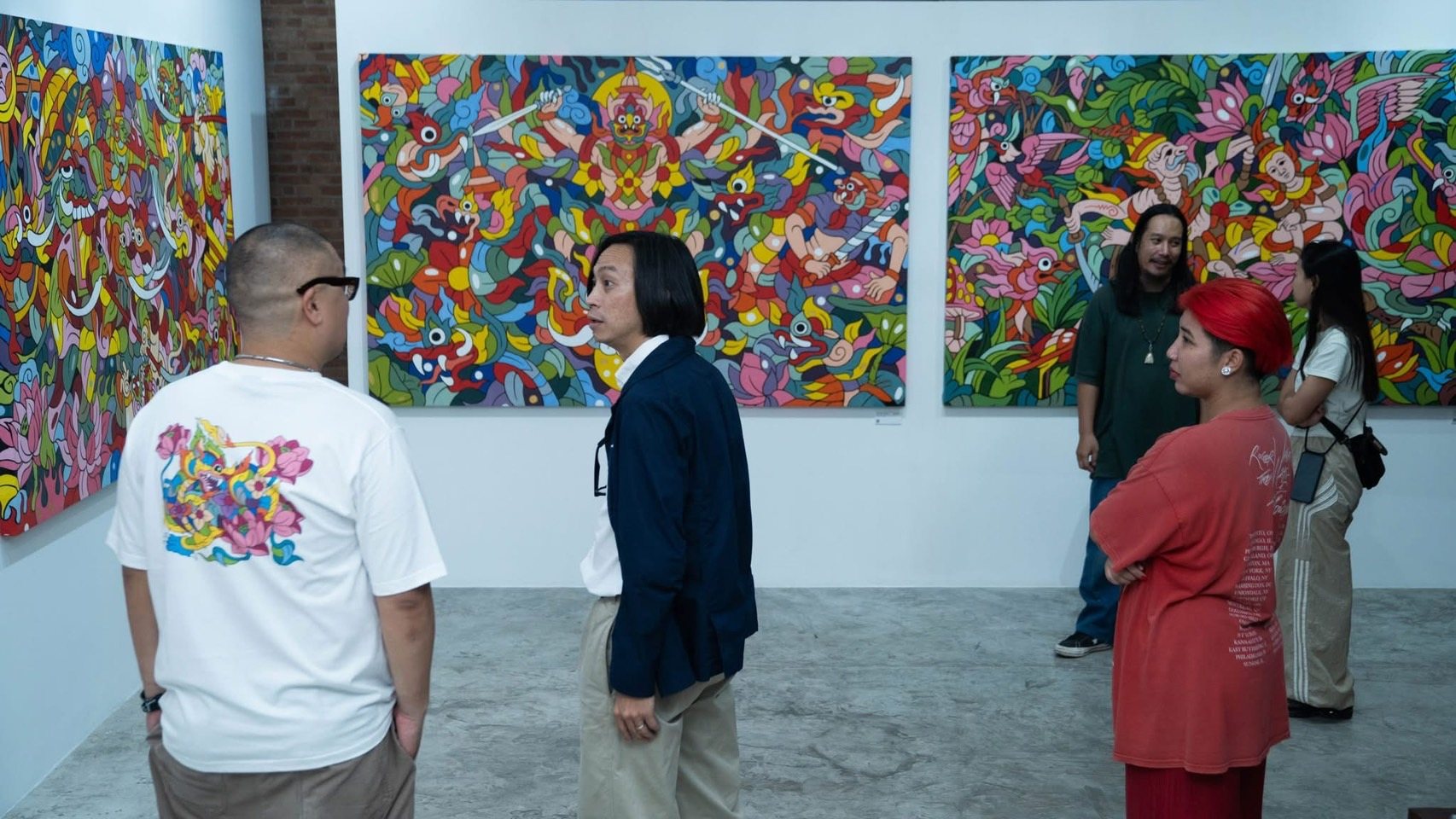 คนห้าคนยืนหน้าภาพสีสดบนผนังสีขาว คนผมแดงกอดอกอยู่ด้านขวา พื้นห้องเป็นสีเทา