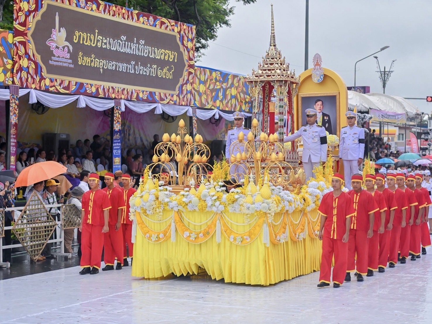 คนชุดแดงเรียงแถวข้างแท่นสีเหลืองประดับดอกไม้ ชายชุดขาวสามคนยืนบนแท่นข้างโครงสร้างยอดแหลม ด้านหลังมีผู้ชมนั่งใต้เต็นท์