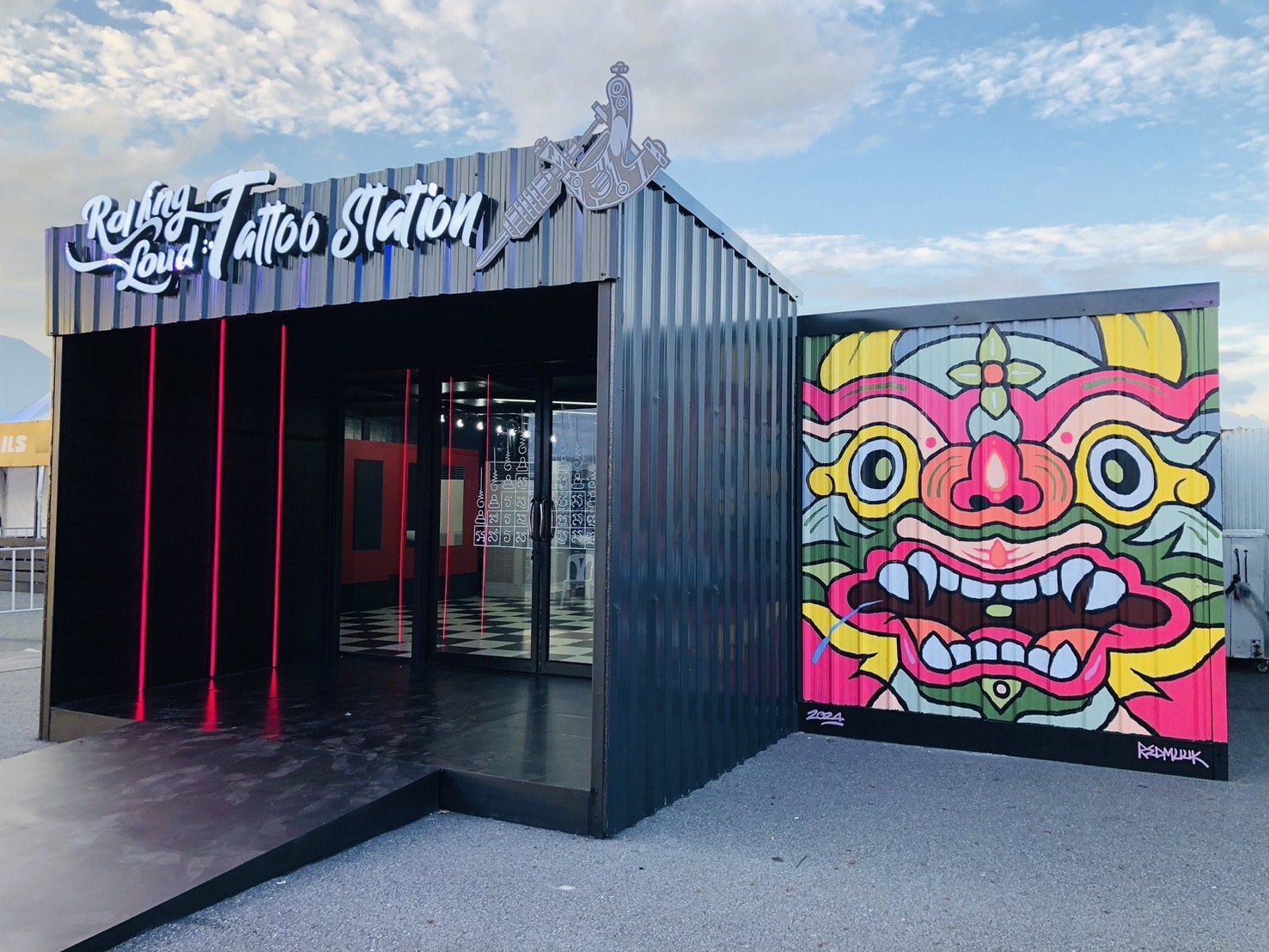 ทางลาดสีดำทอดสู่ประตูกระจก ใต้ป้าย Rolling Loud Tattoo Station ข้างผนังภาพใบหน้าสีชมพู เหลือง เขียว และฟ้า