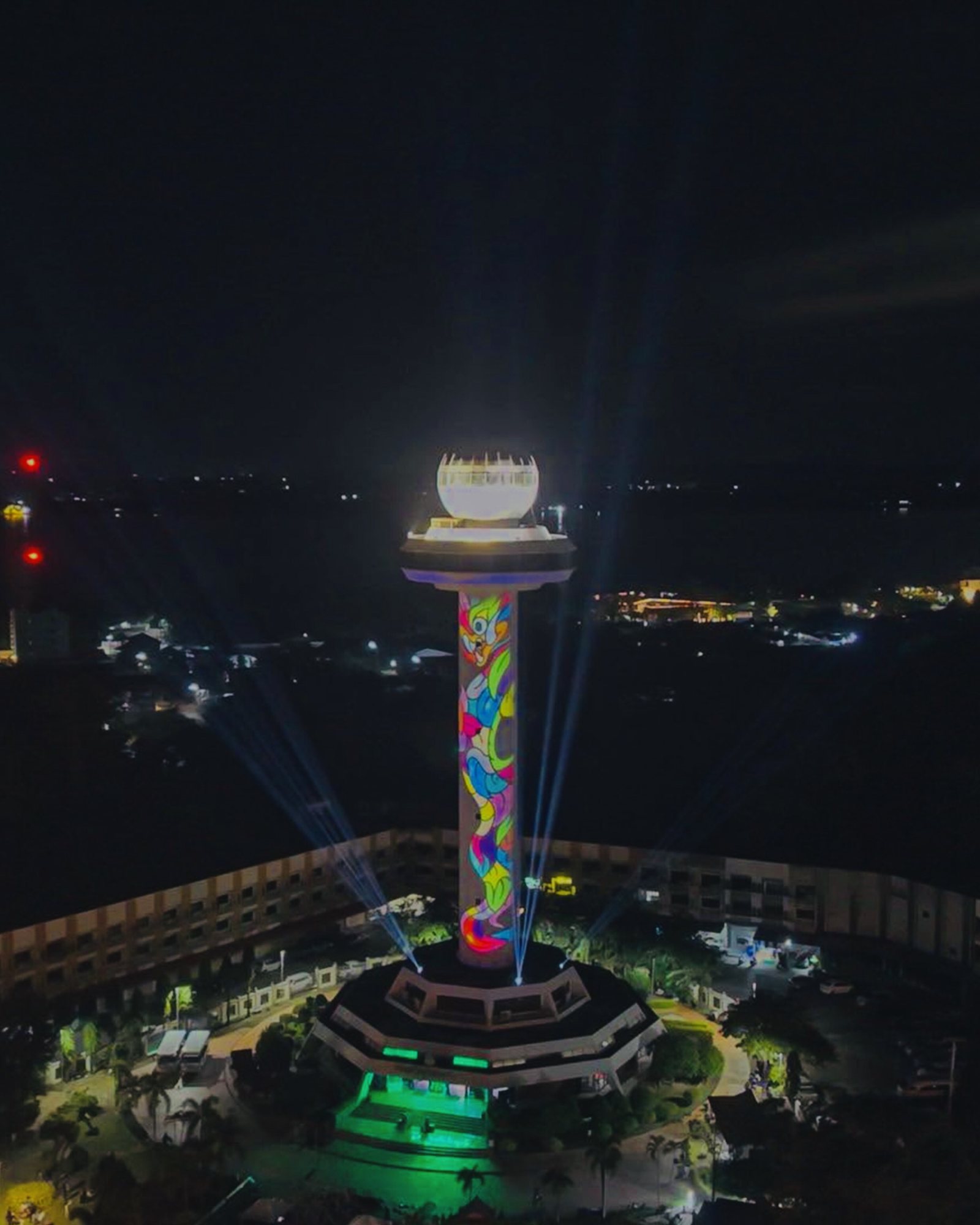 หอคอยลวดลายหลากสียามค่ำคืน ยอดทรงชามส่องสว่าง ลำแสงสีน้ำเงินพุ่งขึ้นรอบฐานอาคารที่มีไฟสีเขียว โดยรอบมีต้นไม้และอาคาร