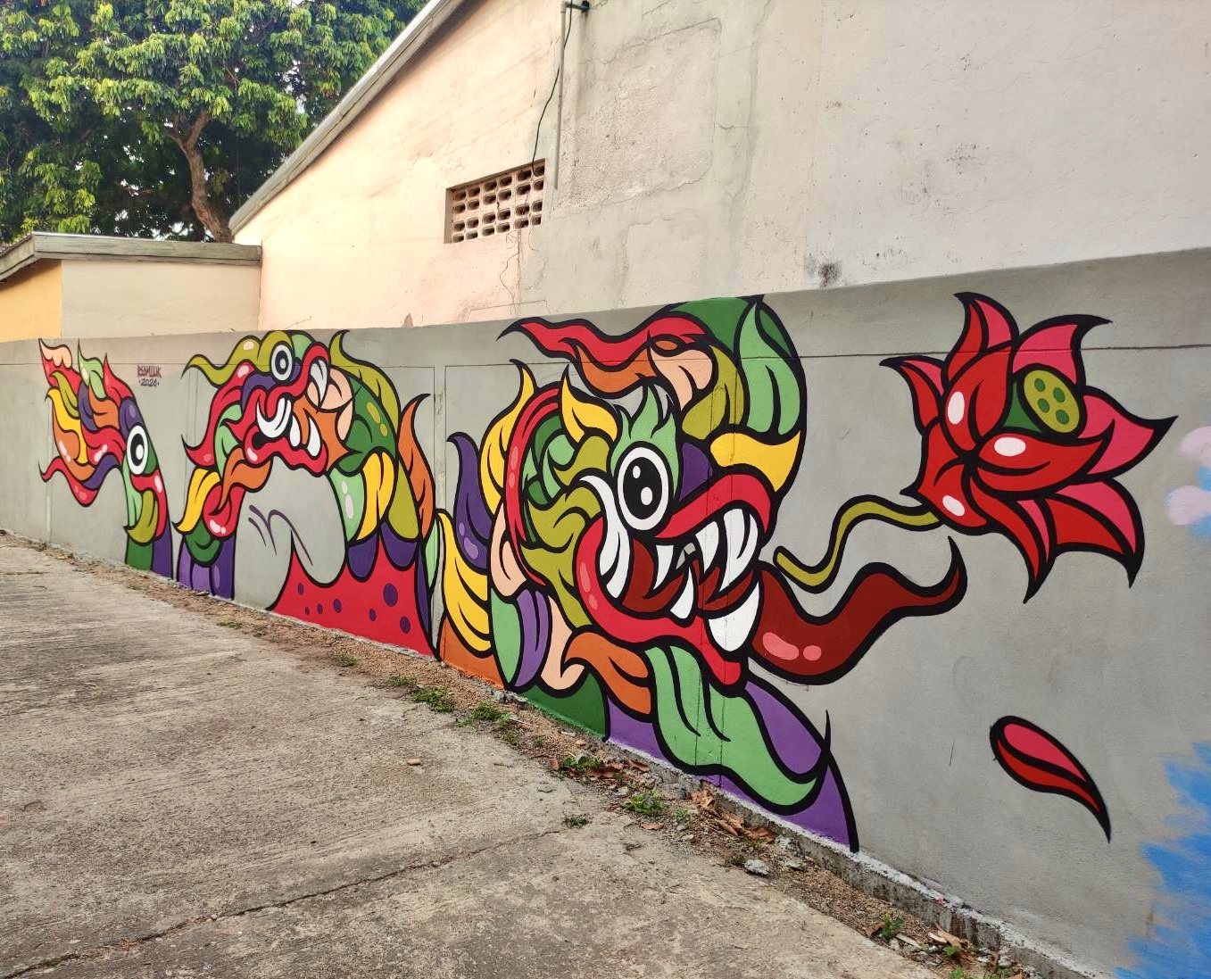 ภาพบนกำแพงสีเทาแสดงสัตว์หลากสี ตาโต เขี้ยวขาว และดอกไม้สีแดง อยู่ข้างทางคอนกรีต มีอาคารและต้นไม้ด้านหลัง