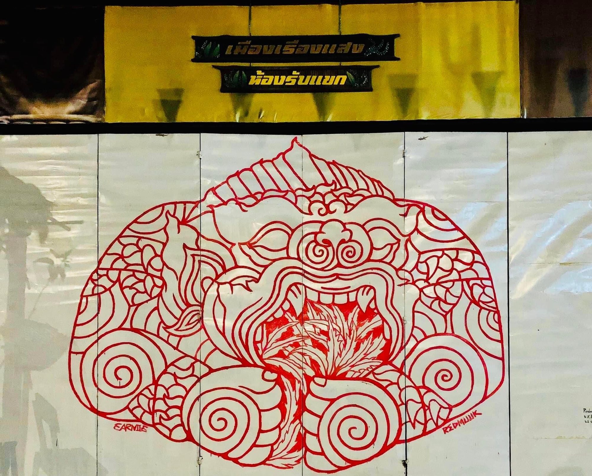 ลายเส้นสีแดงรูปใบหน้ามีเขี้ยวและปากอ้า ล้อมด้วยลายขดและใบไม้ บนผนังสีอ่อน ใต้ป้ายภาษาไทยสองป้าย