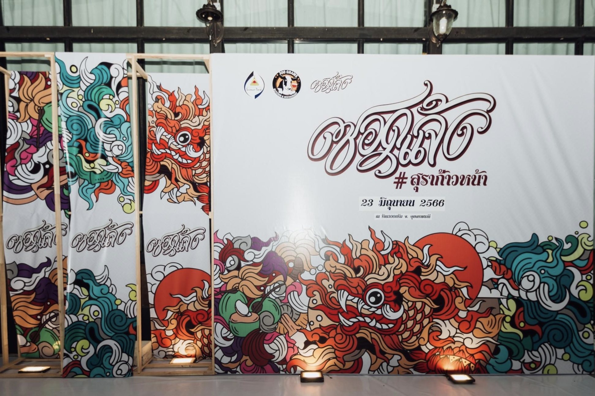 แผงลวดลายใบหน้าหลากสีและตัวอักษรไทย มีกรอบไม้ตั้งด้านซ้าย ไฟบนพื้นส่องขึ้น และโคมไฟสองดวงด้านบน
