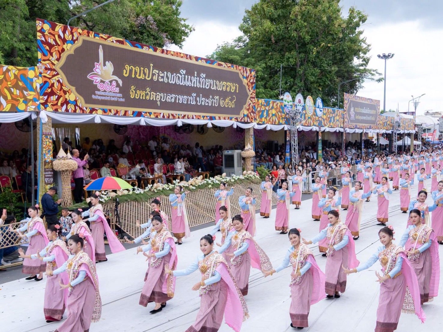 ผู้หญิงสวมชุดสีฟ้าและชมพูเรียงแถวรำบนถนน มีผู้ชมนั่งใต้อัฒจันทร์ตกแต่งลวดลายหลากสีและป้ายอักษรไทย