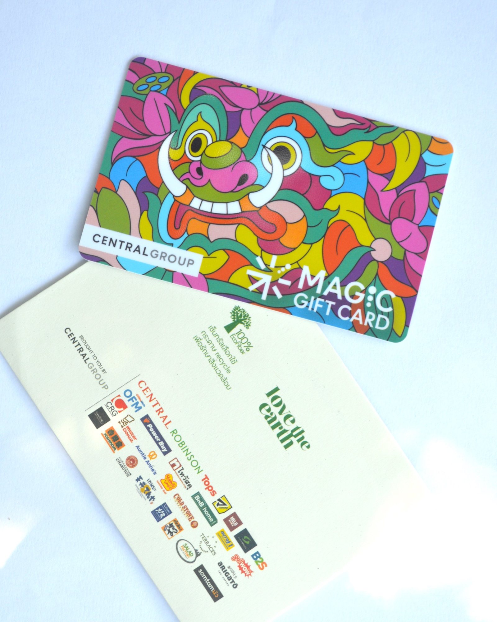 บัตรของขวัญลายใบหน้าสีสด มีเขี้ยวขาวสองข้าง วางเหนือซองสีครีมที่มีข้อความและโลโก้ บนพื้นสีอ่อน