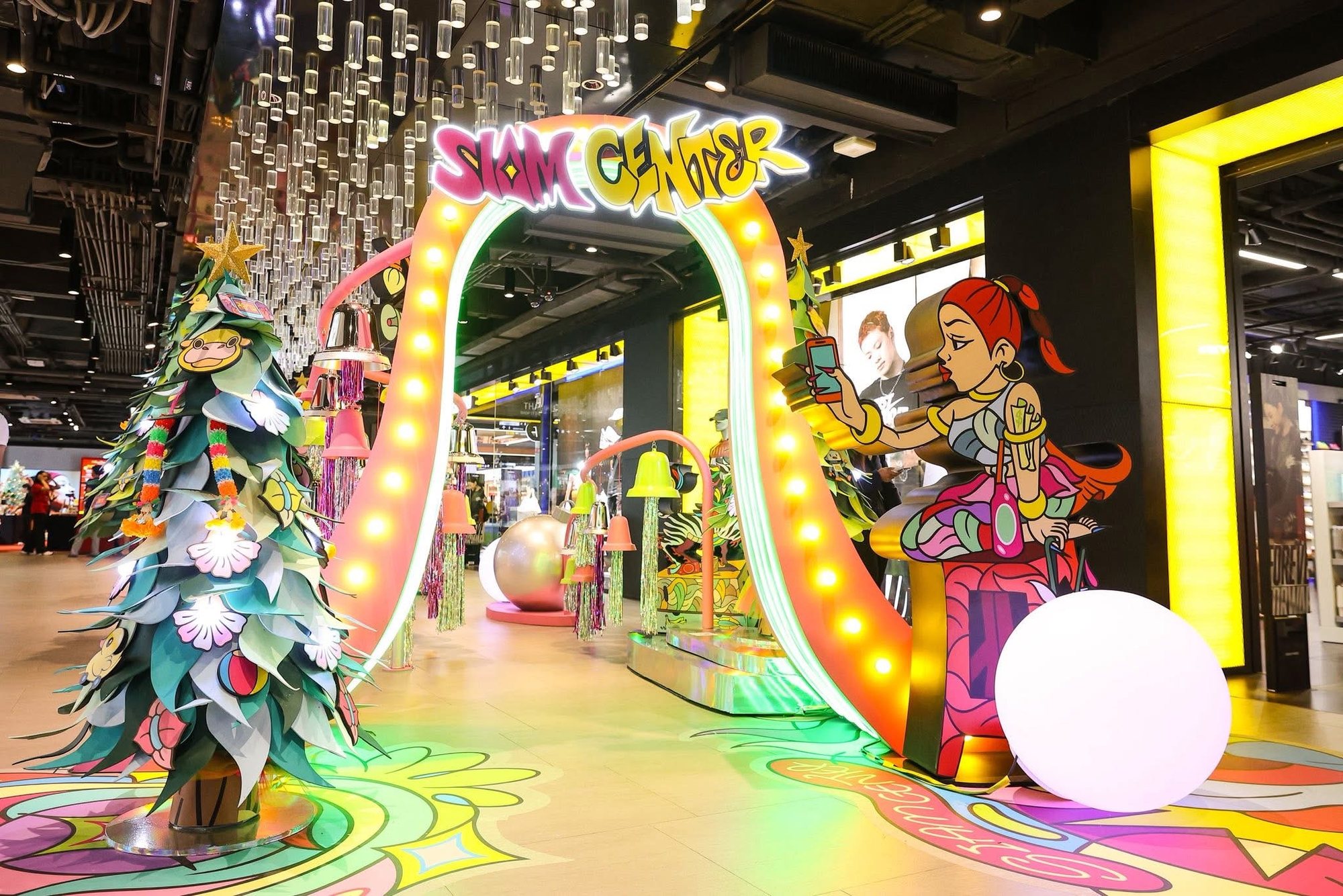 ซุ้มไฟ SIAM CENTER อยู่กลางภาพ ต้นไม้ประดับอยู่ซ้าย รูปการ์ตูนผู้หญิงถือโทรศัพท์และลูกบอลสีขาวอยู่ขวา มีแท่งใสห้อยจากเพดาน
