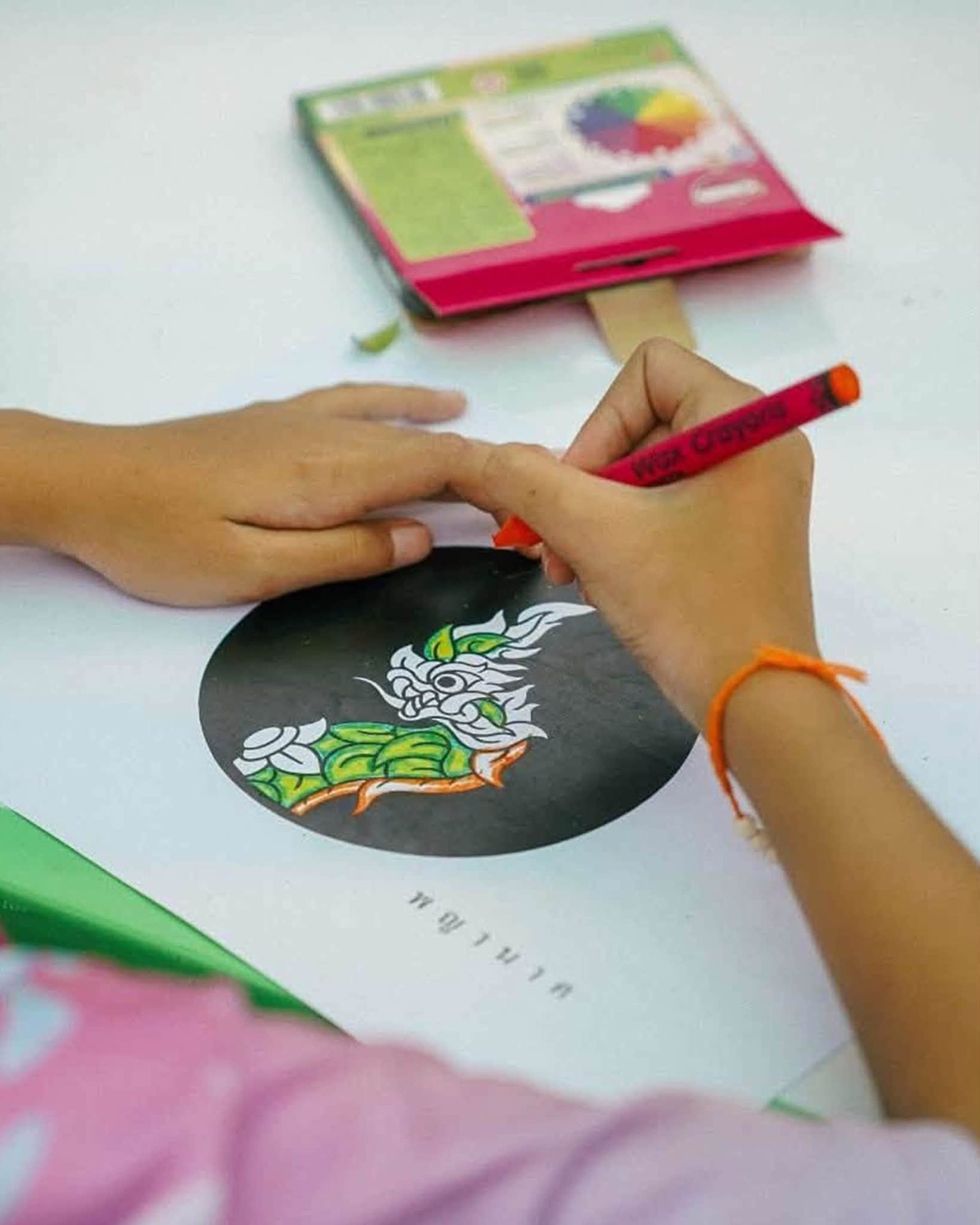 ภาพระยะใกล้ของมือจับสีเทียนด้ามสีแดงเหนือรูปสัตว์ในวงกลมสีดำ มีสายสีส้มผูกข้อมือและกล่องสีด้านหลัง